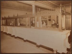 Sepia toned silver print of the interior of First National Bank, circa 1904. Staff can be seen behind the desk and glass partition.